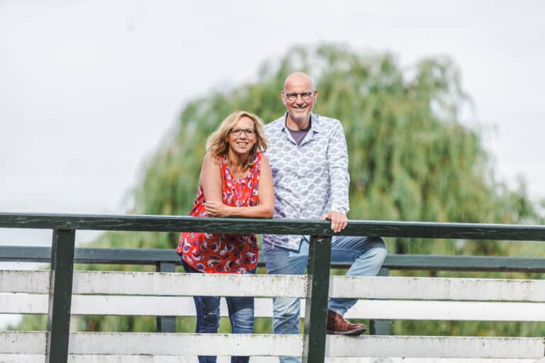 Marco en Jacquelien staan samen op een brug op een mooie zomerse dag en lachen je tegemoet.